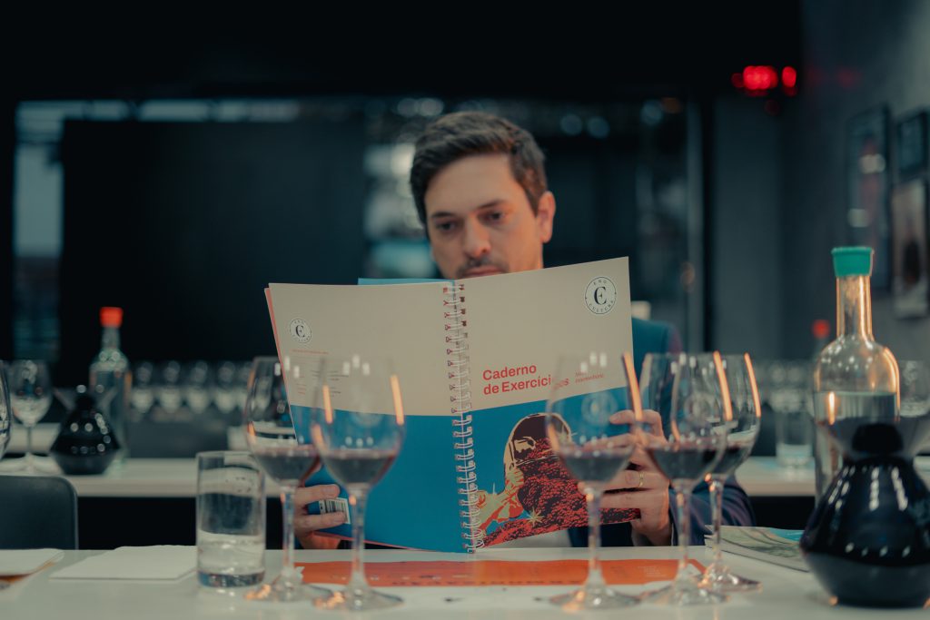 Fotografia em plano médio de um homem concentrado, lendo um material didático da Eno Cultura. Ele segura um caderno de exercícios com espiral, que possui a capa dividida em branco e azul. À sua frente, sobre uma mesa branca, estão alinhadas cinco taças de vinho com bebida tinta, preparadas para uma degustação técnica. O ambiente ao fundo é uma sala de aula ou workshop de vinhos, com iluminação suave e outras taças e garrafas desfocadas.