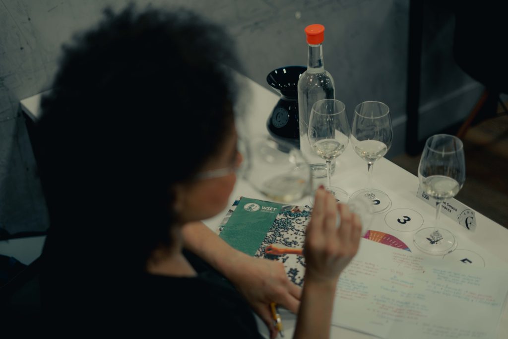 Fotografia em ângulo superior (plongée) mostrando uma mulher de cabelos cacheados durante uma aula de vinhos. Ela segura uma taça de vinho branco contra a luz para análise visual. Sobre a mesa branca, há três taças numeradas, uma garrafa de água, um livro didático da WSET e uma folha de anotações preenchida à mão com caneta. O foco da imagem está nos materiais de estudo e nas taças, com a aluna em primeiro plano levemente desfocada.