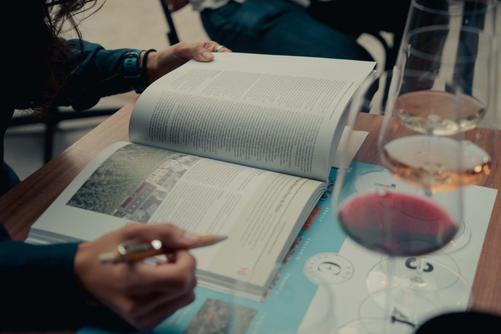 Close das mãos de um aluno folheando uma apostila técnica de vinhos durante uma aula. Ao lado, três taças de cristal com vinhos tinto, rosé e branco alinhadas sobre um guia de degustação da Eno Cultura.