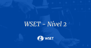 Banner em tons de azul com fundo texturizado em estilo gravura, mostrando uma pessoa servindo vinho em uma taça. No centro, o texto em branco destaca: "WSET – Nível 2". Na parte inferior, centralizado, aparece o logotipo circular da WSET (Wine & Spirit Education Trust).